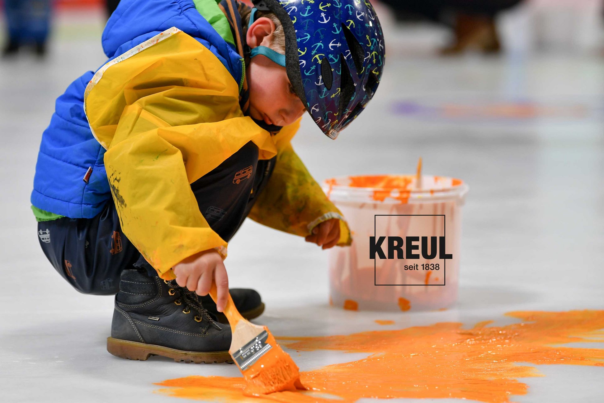 MUCKI Fingerfarbe verdünnt in Eimer für DEG Eismalen auf Eisfläche Eishockey Junge auf Eisfläche malt mit orange farbener Farbe und Pinsel
