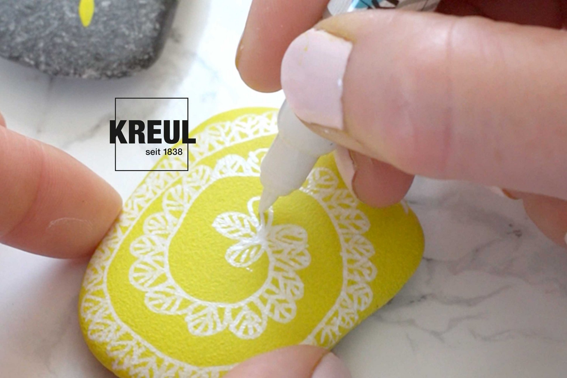 Ein weißer Acrylmarker bemalt einen gelb gefärbten Stein mit einem Linienmuster