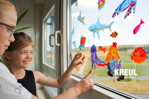 Zwei Mädchen kleben Window Color Motive wie Fische Delfine und eine Meerjungfrau an die Fensterscheibe