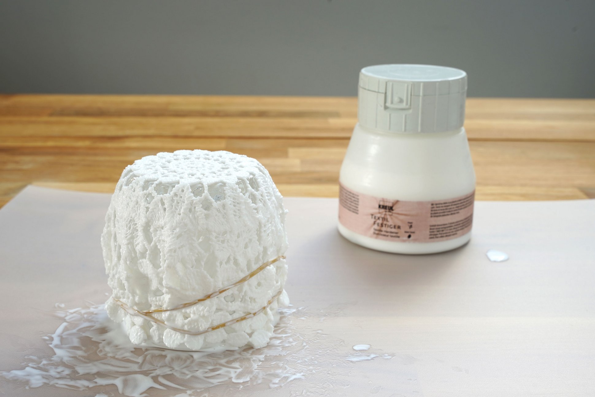 Flasche mit KREUL Textil Festiger hinter einem modellierten, gerundeten Spitzen-Stoff-Stück