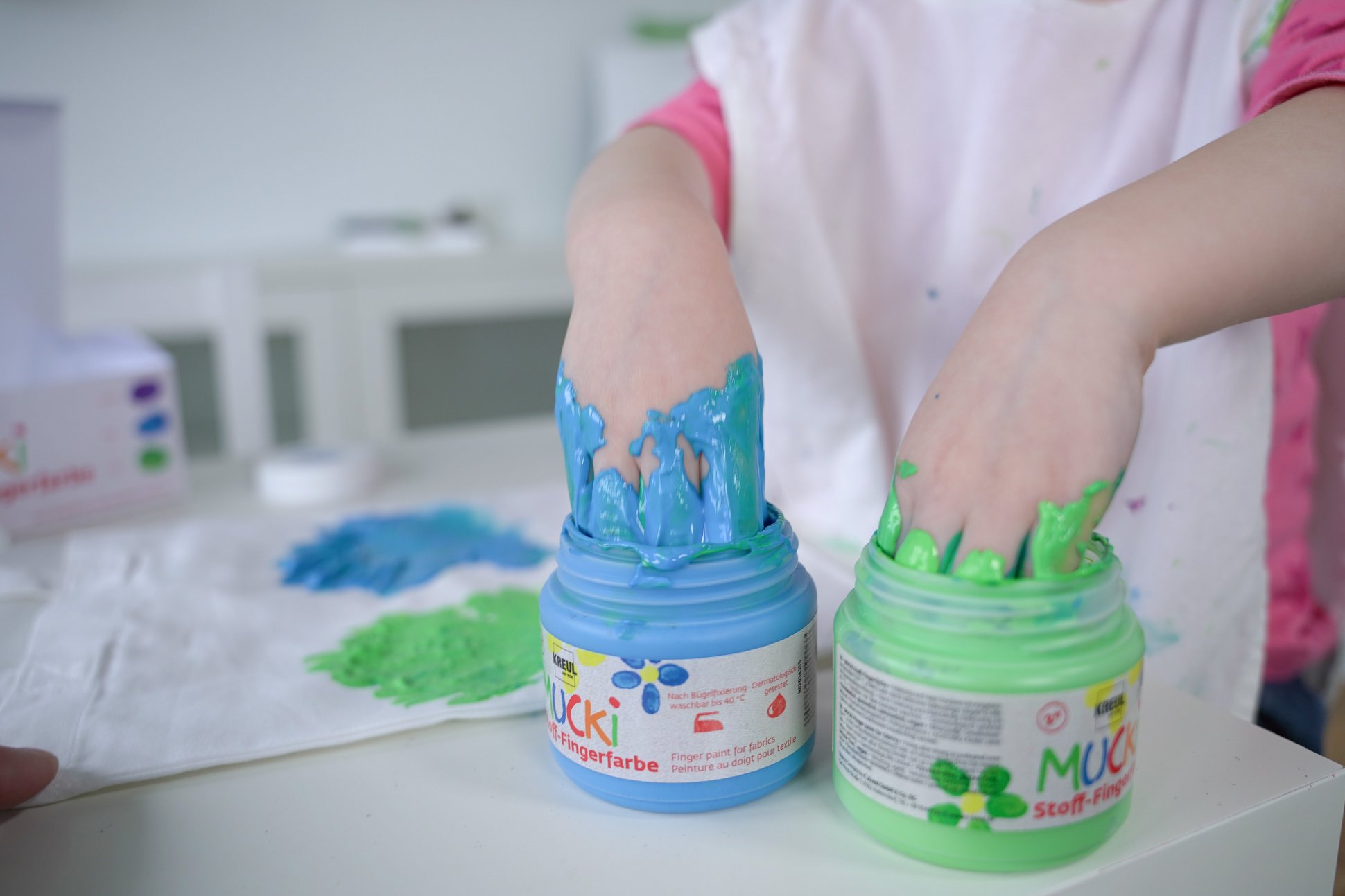 MUCKI Stoff-Fingerfarbe Icon Symbol Bedeutung waschbeständig nach Fixierung Kinderhände tauchen in Dosen mit MUCKI Stoff-Fingerfarbe in Grün und Blau