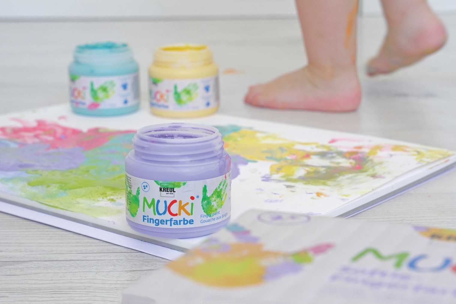 Sicherheit MUCKI Fingerfarbe Dosen mit MUCKI Fingerfarbe in Lila Gelb Hellblau und Grün sowie ein Kinderfuß